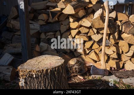 A still-life of a stack of chopped wood and an axe along the side of a wall with the sun shining upon it. Stock Photo