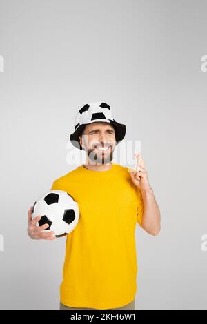 worried football fan in hat holding crossed fingers and soccer ball isolated on grey,stock image Stock Photo