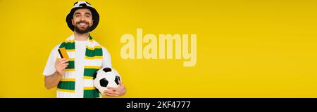 happy man in football fan hat standing with credit card and soccer ball isolated on yellow, banner,stock image Stock Photo