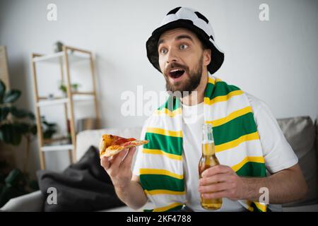 amazed man in hat holding slice of tasty pizza and bottle of beer while watching championship,stock image Stock Photo