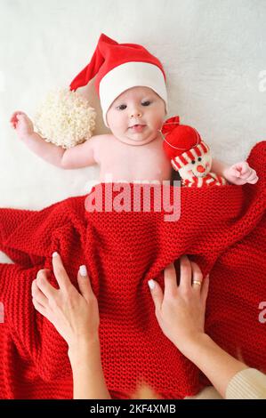 hands cover a baby in a Santa's hat with a red blanket, who holds a snowman toy in his hands. Stock Photo
