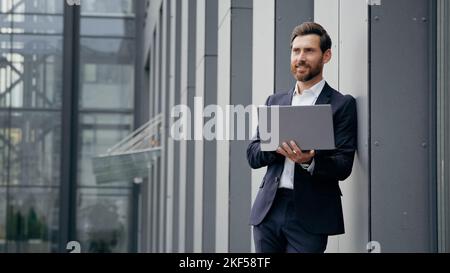 Successful man standing in city near office building typing on laptop professional businessman architect designer develops online project browsing Stock Photo