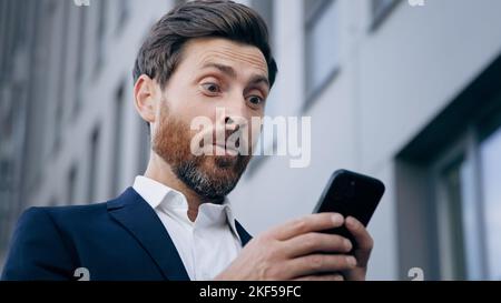 Close-up young surprised man looks at phone screen receiving notification reading good news rejoicing in success satisfied businessman gets excellent Stock Photo