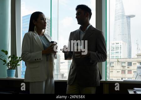 Silhouettes of business people standing at office window, drinking coffee and discussing work Stock Photo