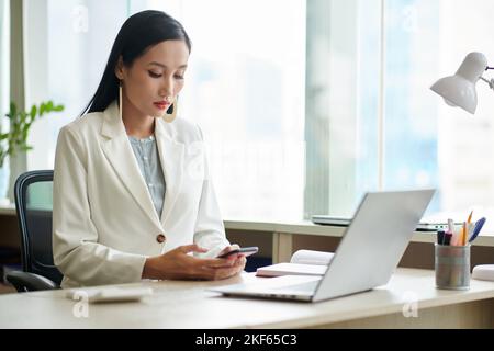 Serious elegant businesswoman working at office desk and checking text messages and notifications on smartphone Stock Photo