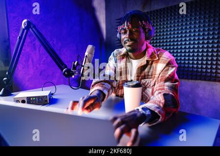African radio host sitting at desk recording in studio with microphone and laptop Stock Photo
