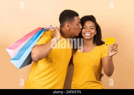 Happy black couple after shopping, woman showing credit card while man holding paper bags and kissing lady Stock Photo