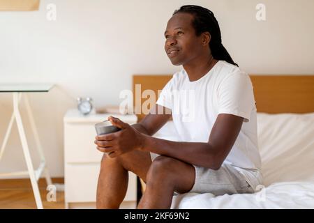 Dreamy Young Black Man Sitting In Bed And Holding Cup Of Coffee Stock Photo