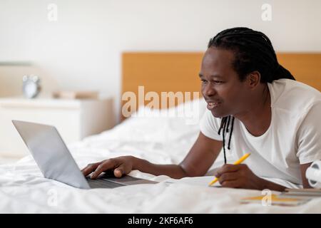 Online Education Concept. Black Young Man Using Laptop While Lying In Bed Stock Photo