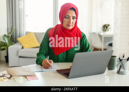 Focused muslim woman in hijab using laptop and calculating budget, sitting at table in living room interior Stock Photo