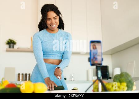 Excited black fit woman in sportswear recording video on smartphone camera, preparing vegetables salad in kitchen Stock Photo
