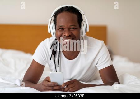 Happy Young Black Man Wearing Wireless Headphones Relaxing In Bed With Smartphone Stock Photo