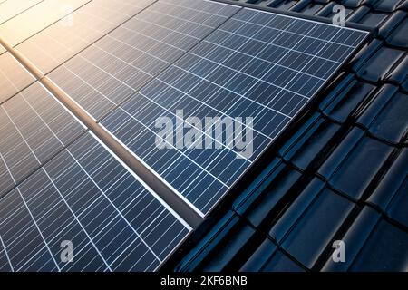 New solar panels are mounted on a roof with shiny black tiles and the clear sky is reflected in the warm sunlight in it. Stock Photo