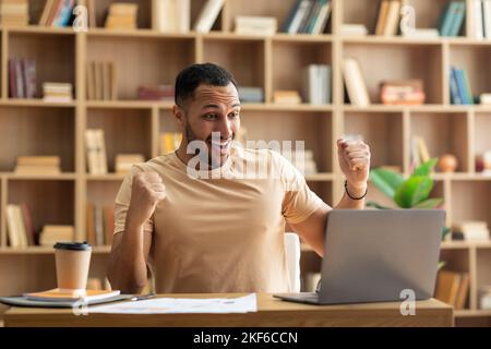 Happy arab male entrepreneur using laptop, shouting and gesturing yes, celebrating business success sitting at workplace Stock Photo