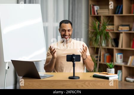 Excited arab man sitting in front of smartphone, recording video or having live videoconference with students Stock Photo