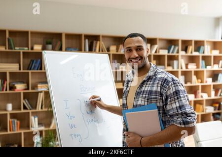 Happy arab male teacher writing rules on whiteboard, looking and smiling at camera in living room interior Stock Photo