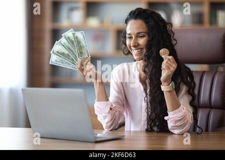 Happy woman sitting in front of laptop, showing bitcoin, cash Stock Photo