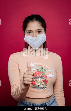 A portrait of a South Asian female wearing a facemask and giving a thumbs up on a red background Stock Photo