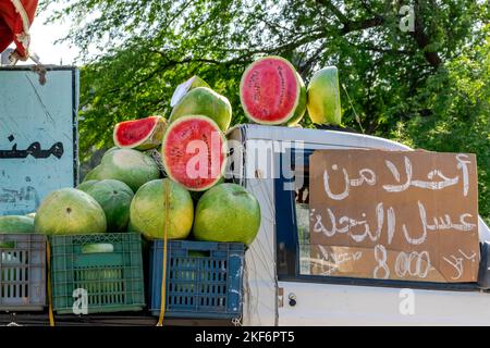 A lot of watermelons for sale. Closed and half opened and piled on crates. Exposed on a mini-truck with an Arabic chalk handwritten board inscription Stock Photo