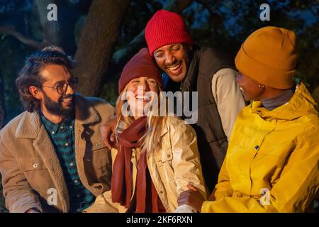 Happy diverse friends have fun laughing next to the bonfire at night caucasian and black couple. Stock Photo