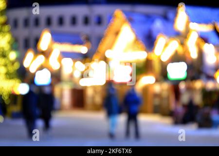 Blurred background. People walk in city square on winter night. Black silhouettes of people walking near houses decorated luminous illumination. White light bokeh blur spots from glowing house lights Stock Photo