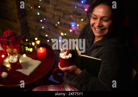 Happy multi-ethnic beautiful woman with cup of hot cocoa with marshmallows, holding a book and smiling a cheerful toothy smile, enjoying magic atmosph Stock Photo