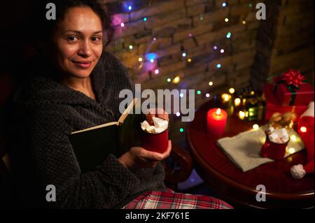 Gorgeous delightful woman with cup of hot chocolate with marshmallows, holding book, smiling a beautiful smile looking at camera, enjoying magic Chris Stock Photo