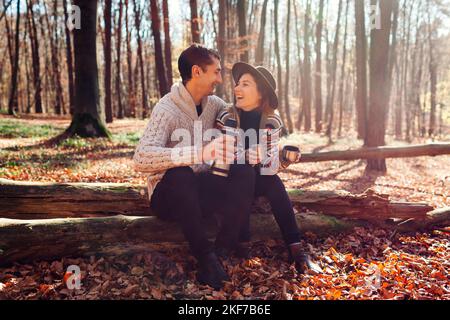 Portrait of young loving couple drinking tea from thermos relaxing in fall forest. Man and woman sit on trunk laughing Stock Photo