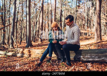 Happy loving couple drinking tea from thermos relaxing in autumn forest. Man and woman sitting on trunk Stock Photo