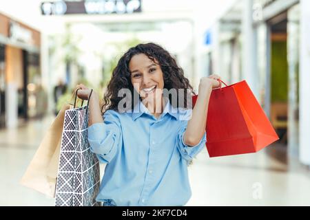 A young Latin American woman tourist bought souvenirs and gifts in another city, country. Standing in the shopping center, holding color packets, lifting and showing them, smiles. Stock Photo