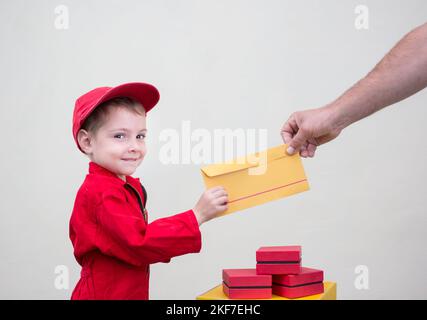 toddler boy in red overalls and cap passes yellow postal envelope to hand of adult. Express transportation, delivery of parcels and letters. postman's Stock Photo