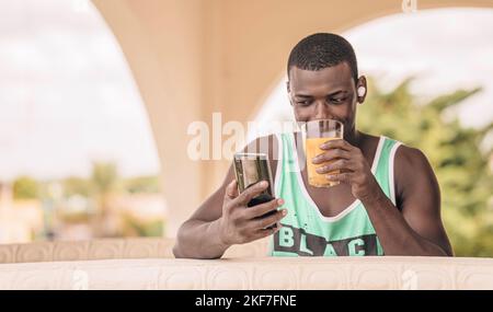 Low angle of black male sipping juice and browsing social media on smartphone while having breakfast on hotel terrace in morning Stock Photo