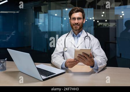 Portrait of a young handsome male doctor who sits and works in an office in a hospital at a table with a laptop. Holds a tablet in his hands, looks at the camera, smiles. Stock Photo