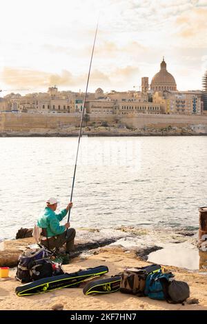 Sliema, Malta - November 12, 2022: Maltese fisherman sitting on a chair on the rocks by the sea while fishing with fishing stick on a sunny morning an Stock Photo