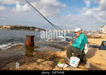 Sliema, Malta - November 12, 2022: Maltese fisherman sitting on a chair on the rocks by the sea while fishing with fishing stick on a sunny morning Stock Photo