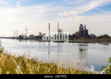 Large steel plant along a river on a sunny summer day. Wind turbines are visible in background. Stock Photo