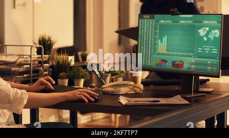 Female employee looking at financial diagrams on monitor, analyzing charts notes and doing paperwork. Working on presentation planning with data, creating startup company report. Stock Photo