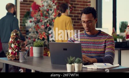 Company employee using laptop in christmas decorated office filled with tree and lights for winter festivity. Young adult working with documents at desk during xmas holiday season. Stock Photo