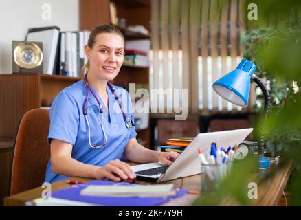 Positive woman doctor working in cabinet on laptop Stock Photo