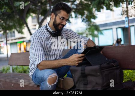 Young bearded man using his tablet sitting on an outdoor bench Stock Photo