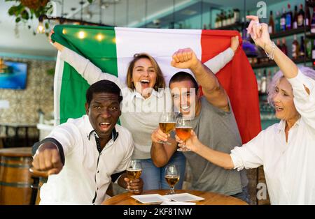 Happy multiracial friends celebrate victory when sports team wins and drinking alcohol drinks in pub, holding flag of Italy Stock Photo