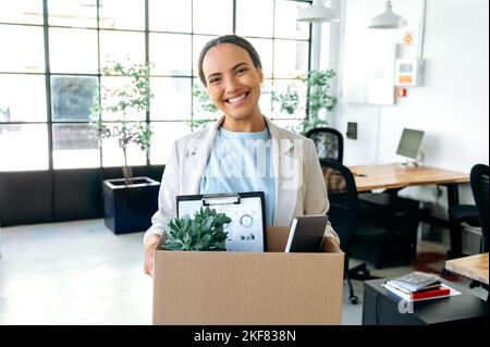 Happy joyful brazilian or hispanic curly woman, in elegant clothes, trainee, financial manager, got a new job, standing in the office, holding a box with office supplies, looks at camera, smiling Stock Photo