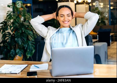 Satisfied relaxed mixed race female employee resting during work time, relaxed sits at her workplace, puts hands behind head, feels satisfied by project done, looking at laptop, smiling friendly Stock Photo