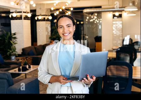 Photo of a confident beautiful elegant young mixed race woman, successful company worker, seo, programmer, standing in her office, holds an open laptop, looking at the camera, smiling friendly Stock Photo