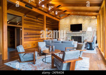 Brown wooden and grey upholstered sitting chairs, round table and sofas with natural stone fireplace in living room inside cedar and timber home. Stock Photo