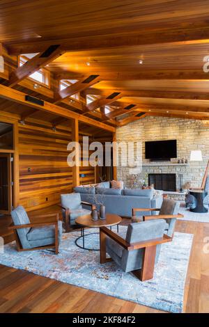 Brown wooden and grey upholstered sitting chairs, round table and sofas with natural stone fireplace in living room inside cedar and timber home. Stock Photo