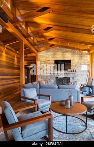 Brown wooden and grey upholstered sitting chairs, round table and sofas in living room area of great room inside luxurious cedar and timber wood home. Stock Photo