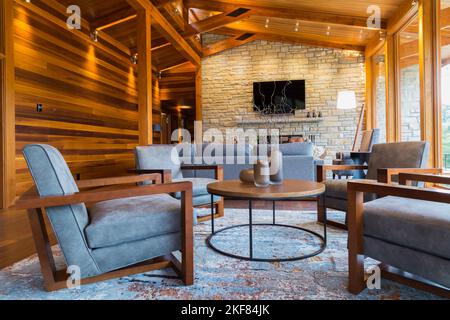 Brown wooden and grey upholstered sitting chairs, round table and sofas in living room area of great room inside luxurious cedar and timber wood home. Stock Photo