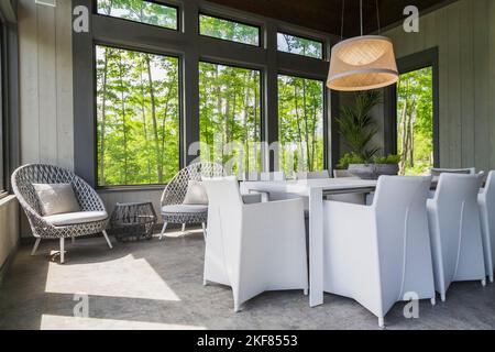 White dining table with white linen upholstered armchairs plus grey wicker chairs in gazebo inside a luxurious contemporary bungalow style home. Stock Photo