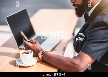 A bearded black African businessman in a street cafe, with a selective focus on his hand typing a message on the cellphone; a cup of espresso coffee n Stock Photo